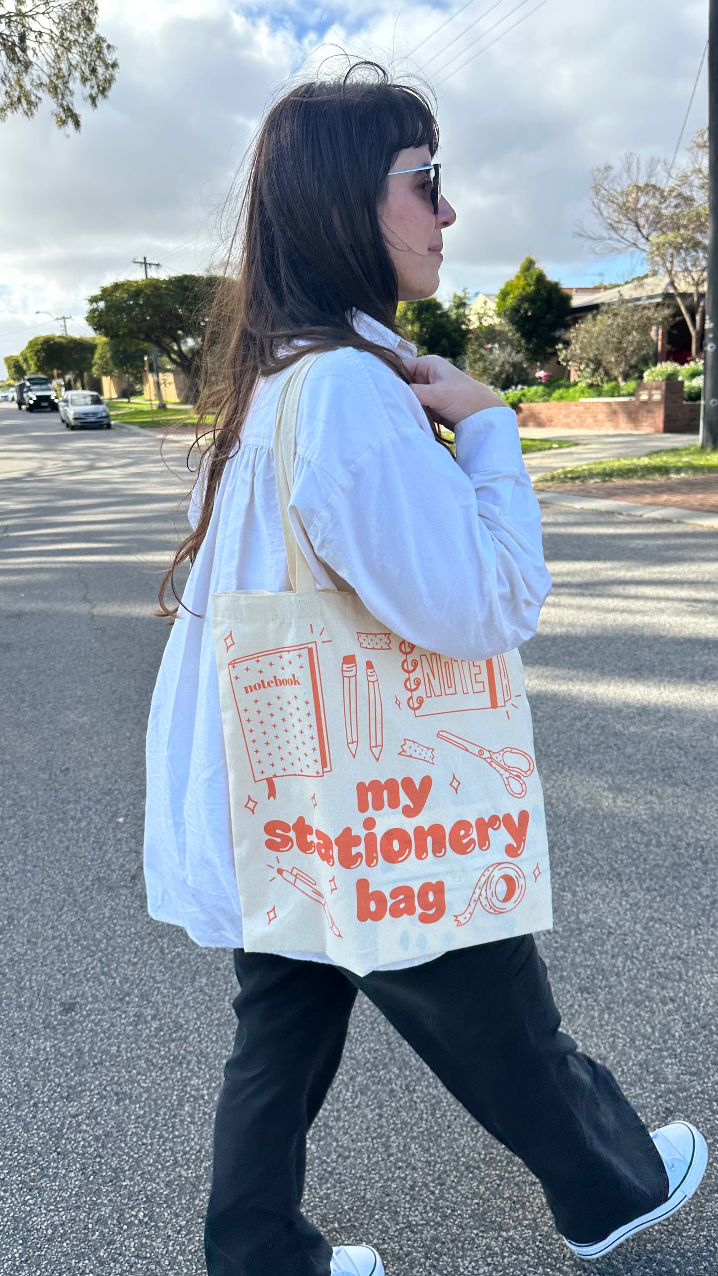 A woman walking outdoors on a street wearing a white button-up shirt, black pants, and white sneakers, carrying a cream canvas tote bag with orange text reading 'my stationery bag' and stationery illustrations.
