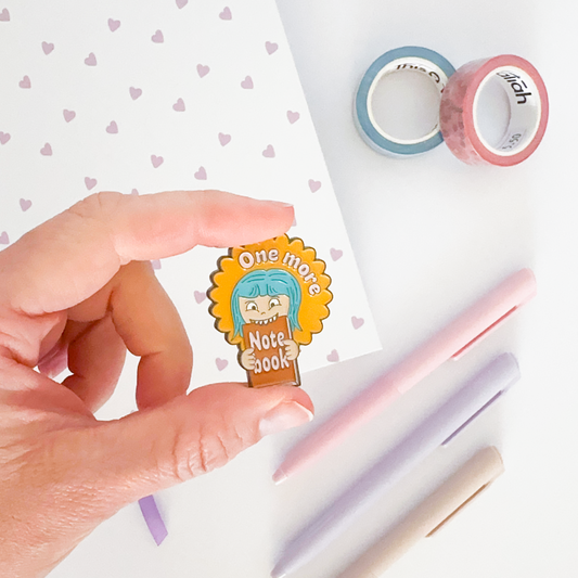 Enamel pin with notebook, coloured pen and washi tapes in the background