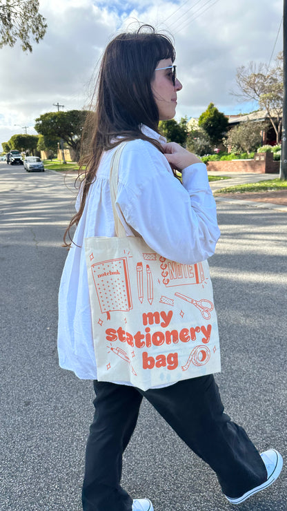 A woman walking outdoors on a street wearing a white button-up shirt, black pants, and white sneakers, carrying a cream canvas tote bag with orange text reading 'my stationery bag' and stationery illustrations.