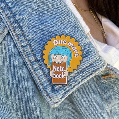 A close-up of a person wearing a light blue denim jacket over a white shirt, featuring a cartoon enamel pin on the lapel. The pin shows a blue-haired character holding a notebook with the text 'One more Note book'.