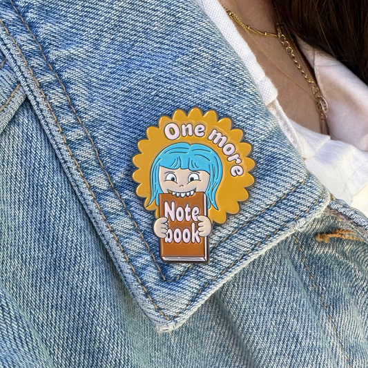 A close-up of a person wearing a light blue denim jacket over a white shirt, featuring a cartoon enamel pin on the lapel. The pin shows a blue-haired character holding a notebook with the text 'One more Note book'.