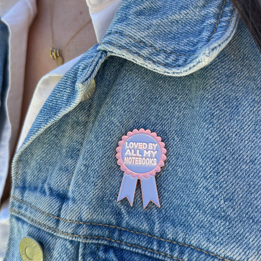 A close-up of a person wearing a blue denim jacket over a white collared shirt, featuring a pink and lavender enamel pin on the jacket that reads 'LOVED BY ALL MY KITSURING'.