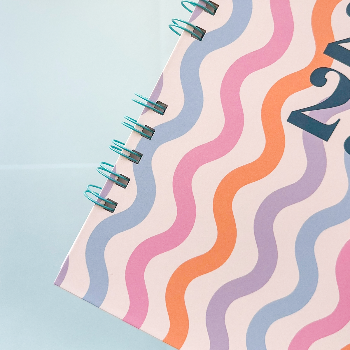 A close-up of a spiral-bound notebook or planner page with a pastel wavy stripe pattern in pink, orange, lavender, and blue on a pale background, bound with a turquoise wire coil.
