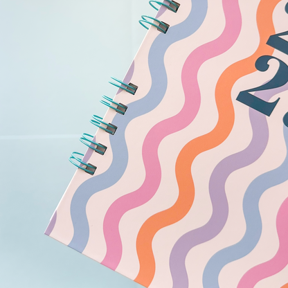 A close-up of a spiral-bound notebook or planner page with a pastel wavy stripe pattern in pink, orange, lavender, and blue on a pale background, bound with a turquoise wire coil.