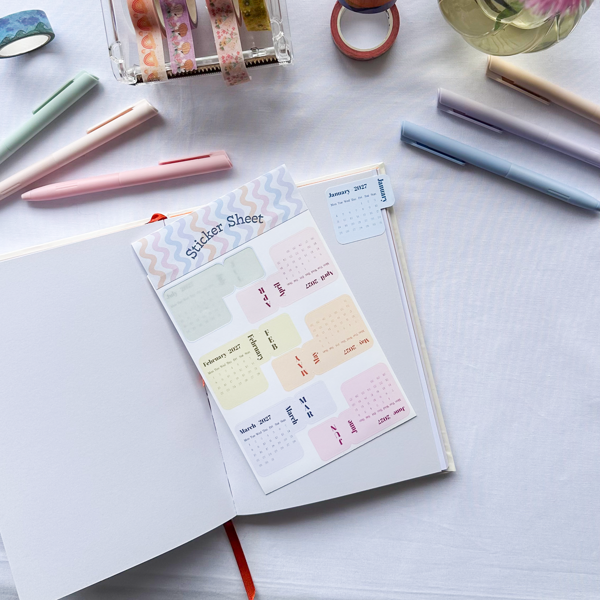 A styled flat-lay of a planner sticker sheet labeled 'Gadget Sheet' placed on an open notebook, surrounded by pens, washi tape, and flowers on a white fabric background.