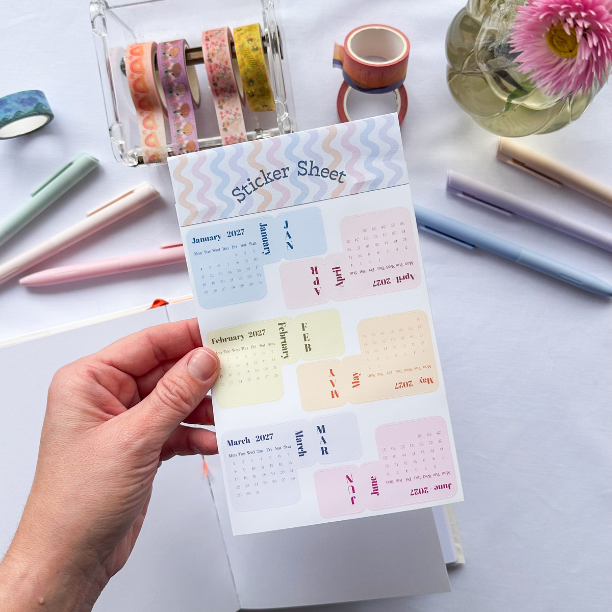A hand holds a planner sticker sheet with pastel monthly calendar stickers and decorative chevron borders, photographed on a white tabletop with pens, washi tape, and flowers in the background.