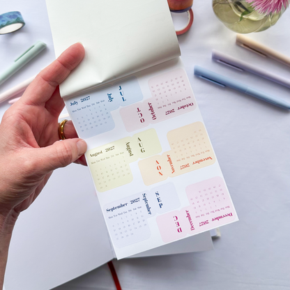 A hand holds an open planner sticker sheet with pastel monthly calendar stickers, shown on a white desk with pens, washi tape, and pink flowers in the background.