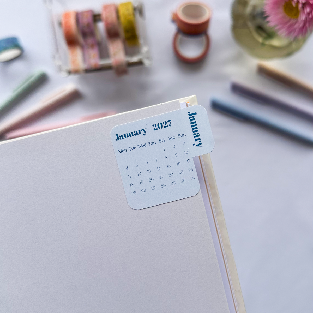 A close-up of a white planner or notebook page with a small sticker showing January 2027, styled on a light surface with pens, washi tape, and pink flowers in the background.