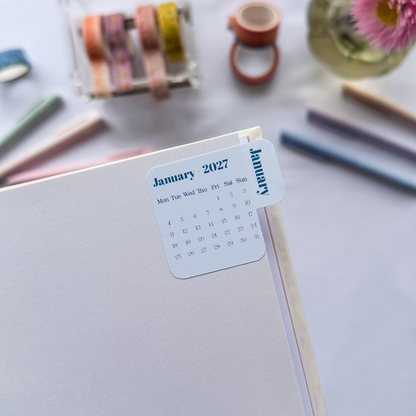 A close-up of a white planner or notebook page with a small sticker showing January 2027, styled on a light surface with pens, washi tape, and pink flowers in the background.