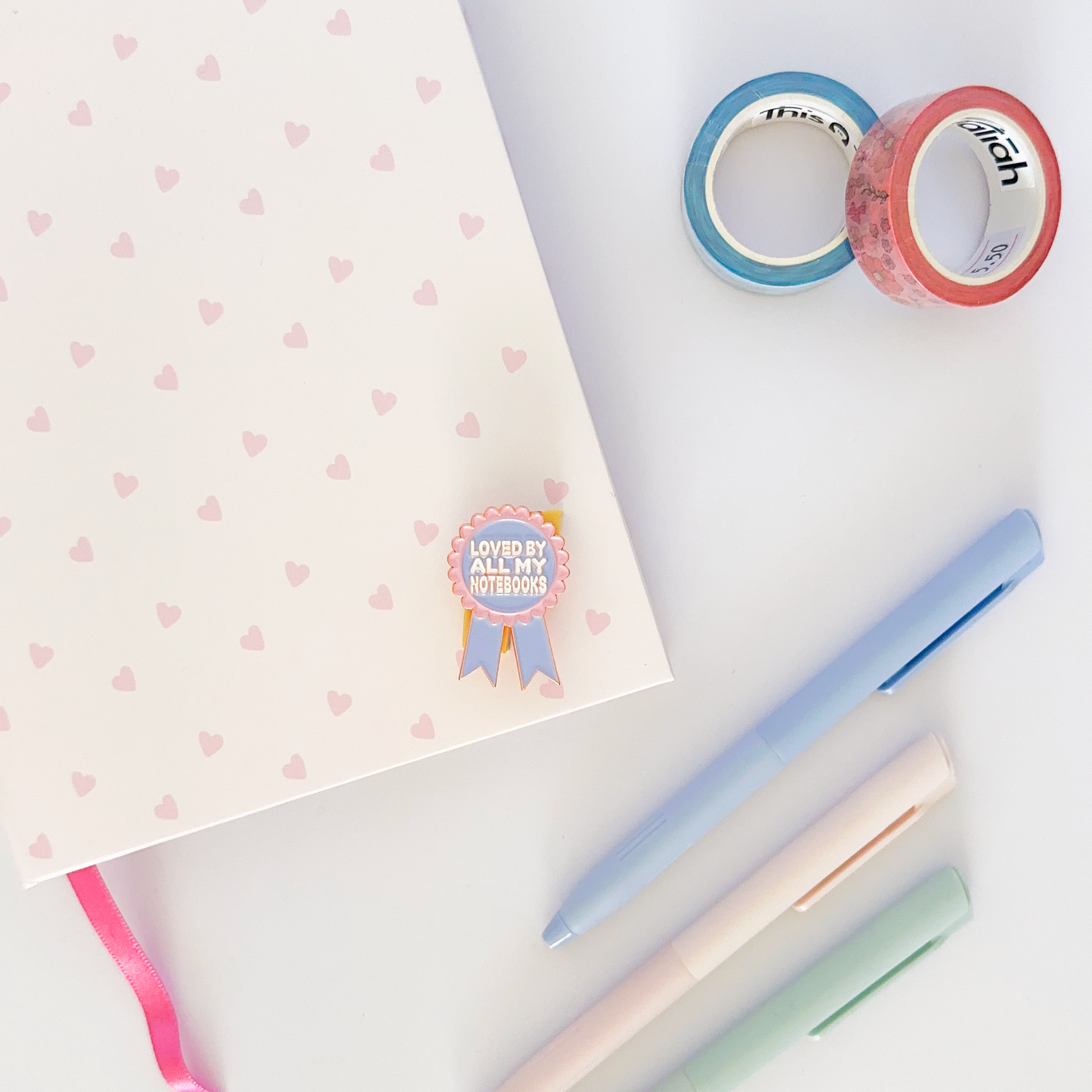 Pink and purple enamel pin with notebook, coloured pen and washi tapes in the background.