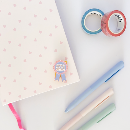 Pink and purple enamel pin with notebook, coloured pen and washi tapes in the background.