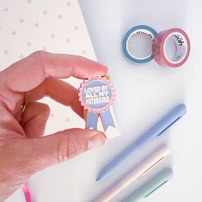 Pink and purple enamel pin with notebook, coloured pen and washi tapes in the background.