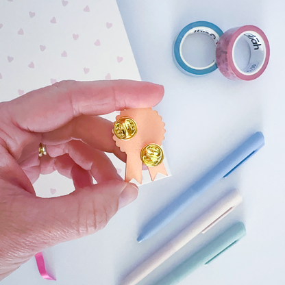 Pink and purple enamel pin with notebook, coloured pen and washi tapes in the background. 