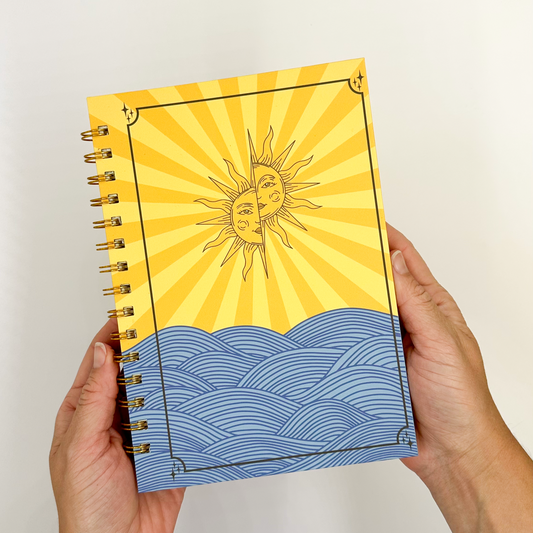 Notebook with sun and wave design held by hands against a plain background