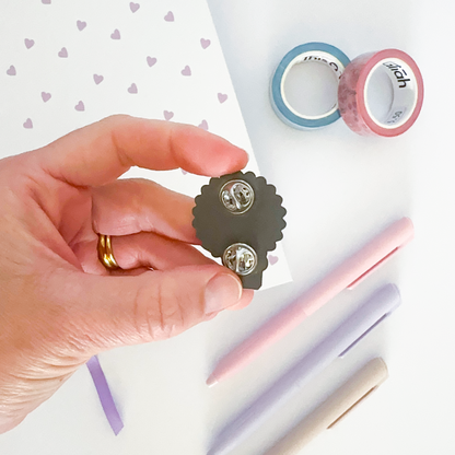 Enamel pin with notebook, coloured pen and washi tapes in the background