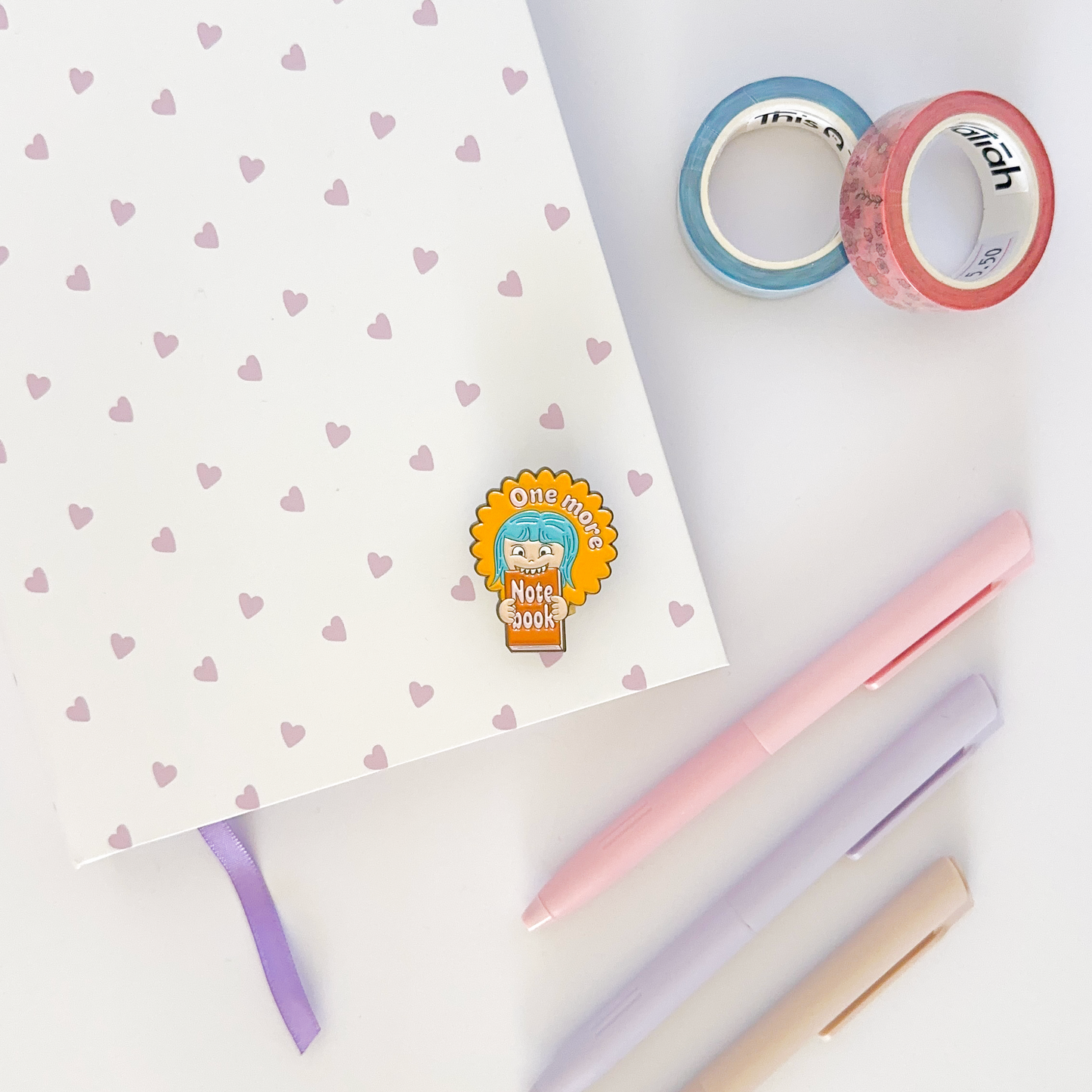 Enamel pin with notebook, coloured pen and washi tapes in the background