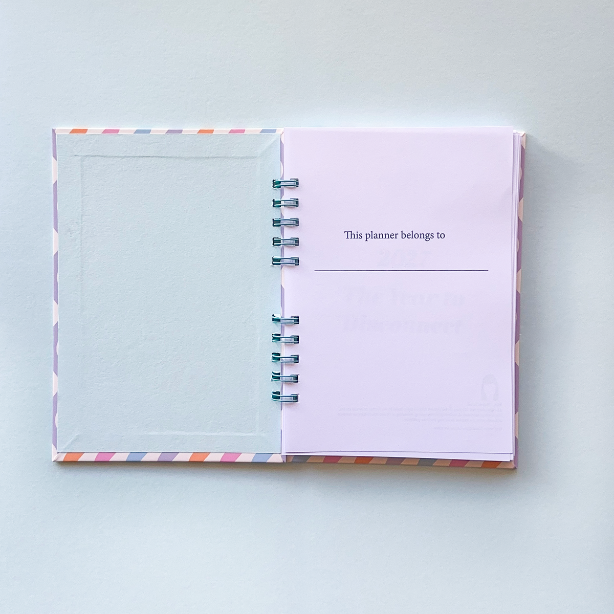An open spiral-bound planner with a pastel lavender page that reads 'This planner belongs to:' and a blank lined space below, photographed on a light gray background.