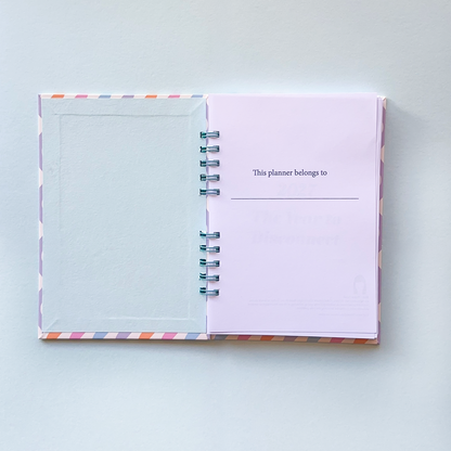 An open spiral-bound planner with a pastel lavender page that reads 'This planner belongs to:' and a blank lined space below, photographed on a light gray background.