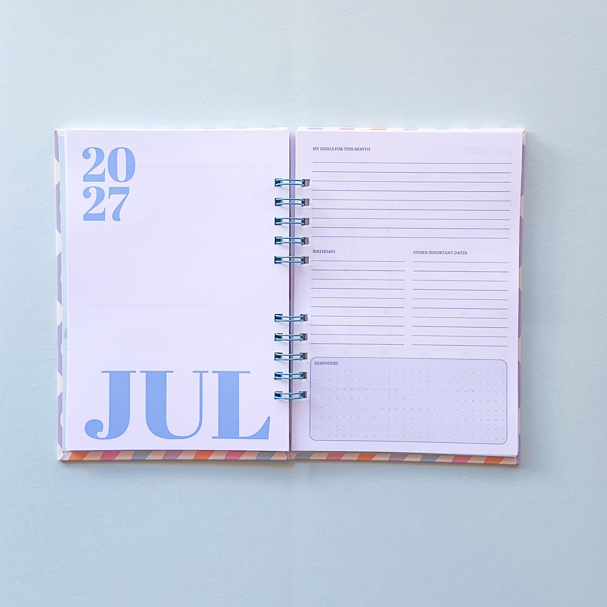 An open spiral-bound planner showing July 2027, with lined daily planning pages and a pastel multicolor patterned cover, photographed on a light blue background.