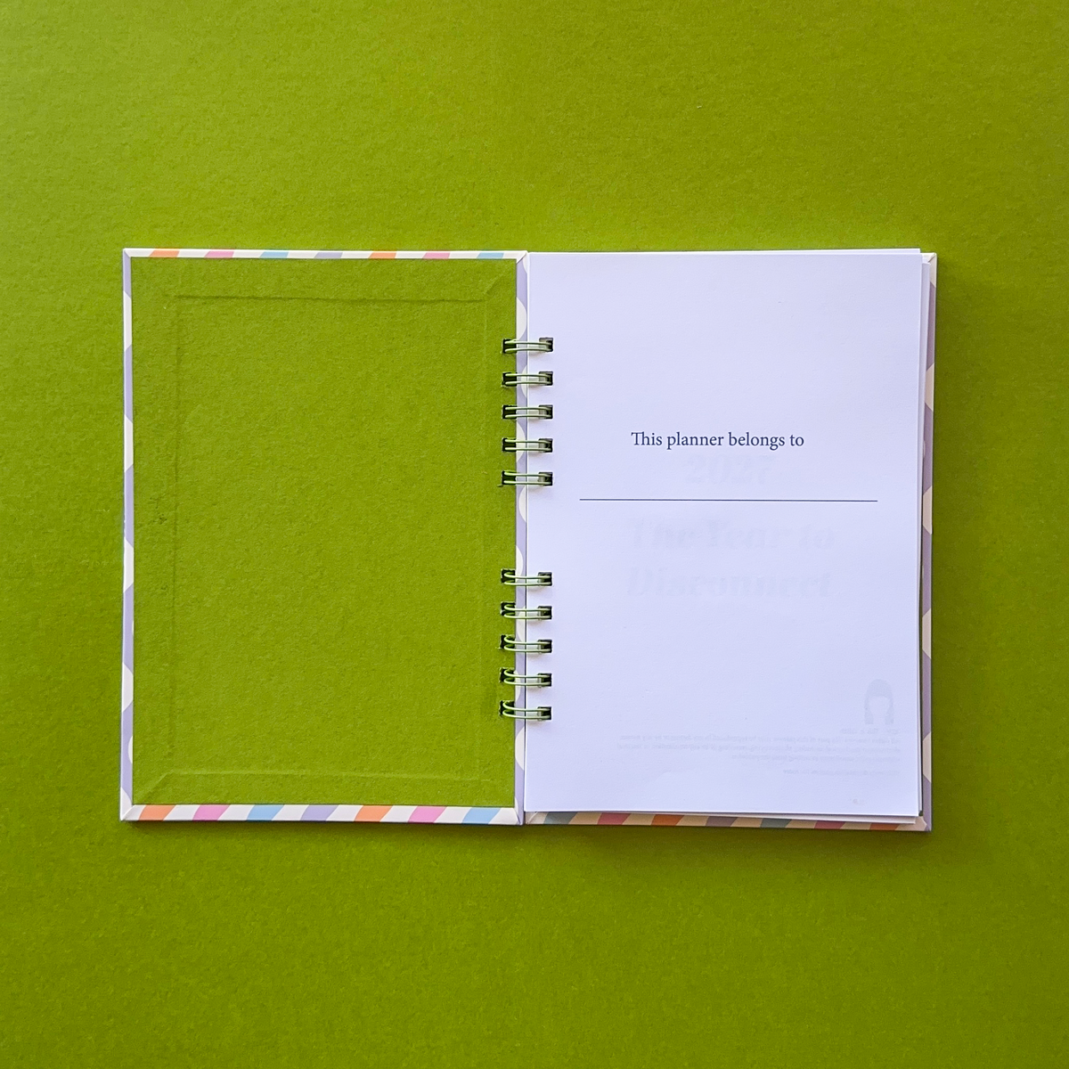 An open spiral-bound planner or journal lies on a solid olive green background. The left page is a solid green cover with a thin multicolor striped border, and the right page is white with printed text that reads 'This planner belongs to'.
