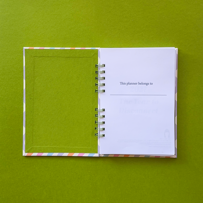An open spiral-bound planner or journal lies on a solid olive green background. The left page is a solid green cover with a thin multicolor striped border, and the right page is white with printed text that reads 'This planner belongs to'.