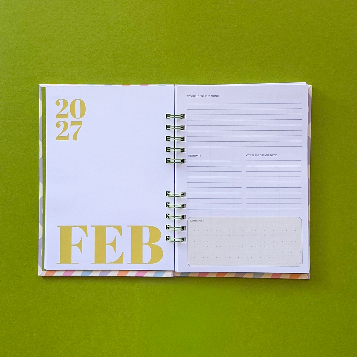An open spiral-bound planner or calendar showing February 2027, with white pages and green text, photographed on a solid olive green background.