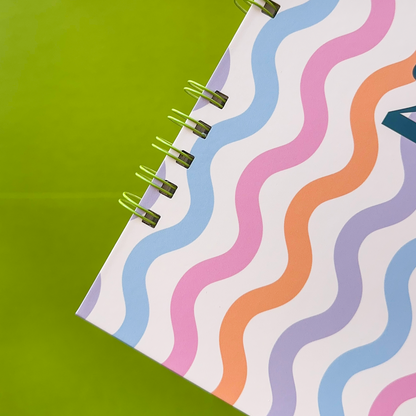 A close-up of a spiral-bound notebook with a white cover decorated with wavy pastel stripes in pink, blue, orange, and purple, set against a bright green background and green wire bind.