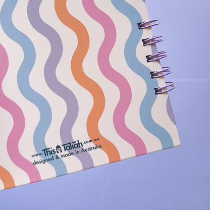A spiral-bound notebook with a white cover featuring retro wavy stripes in pastel pink, orange, purple, and blue, photographed on a light blue background.