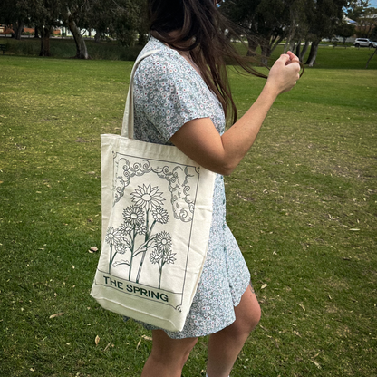 A woman in a light blue floral dress stands in a grassy park, carrying a cream canvas tote bag with a black line-art floral design and the text 'THE SPRING'.