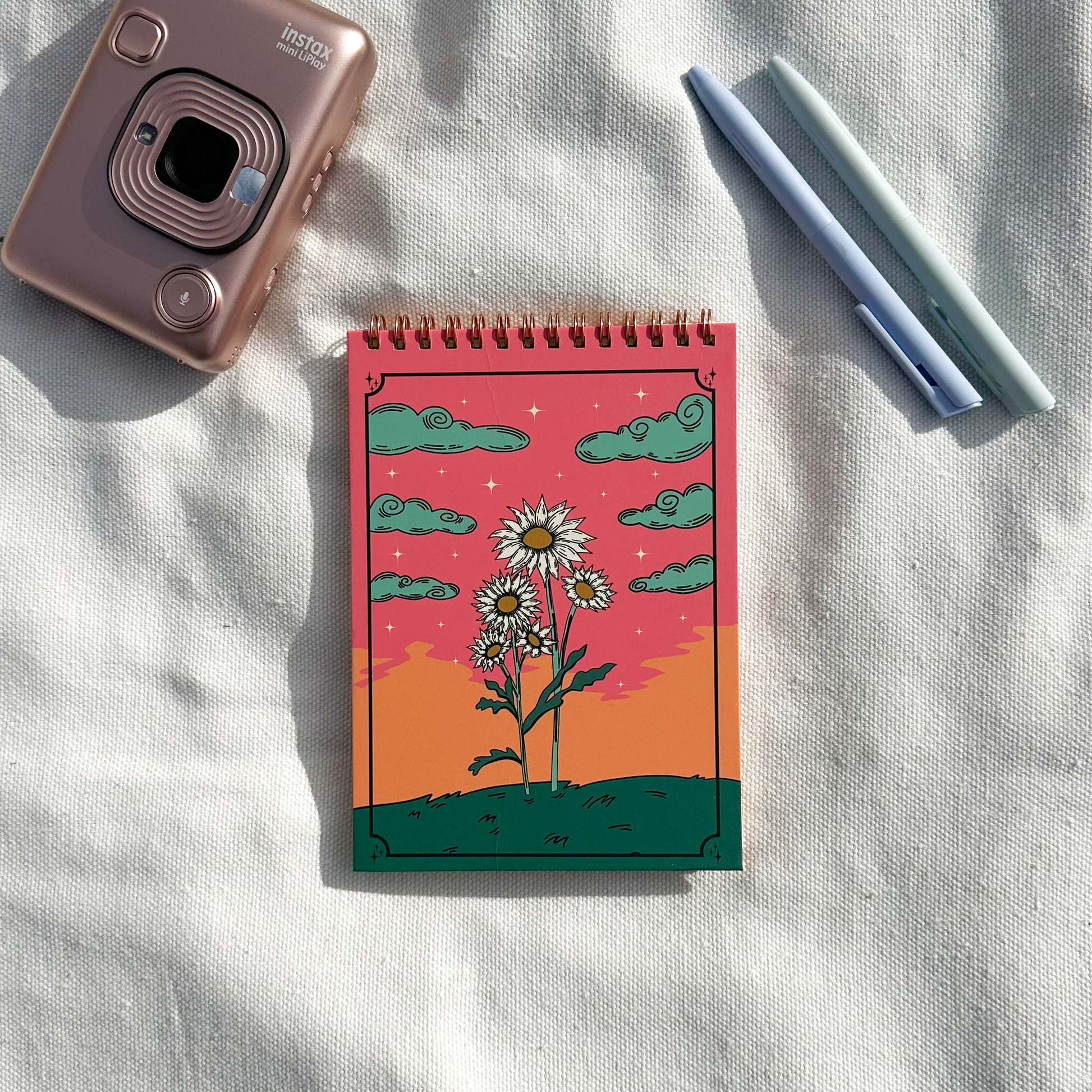 Spring notepad showing flower design with pink sky with green grass and green clouds in a light beige towel and polaroid and pens