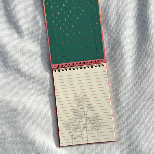 A small spiral-bound notebook lies open on a white fabric surface. The cover is dark green with a repeating small gold motif, and the lined interior shows a faint tree illustration.