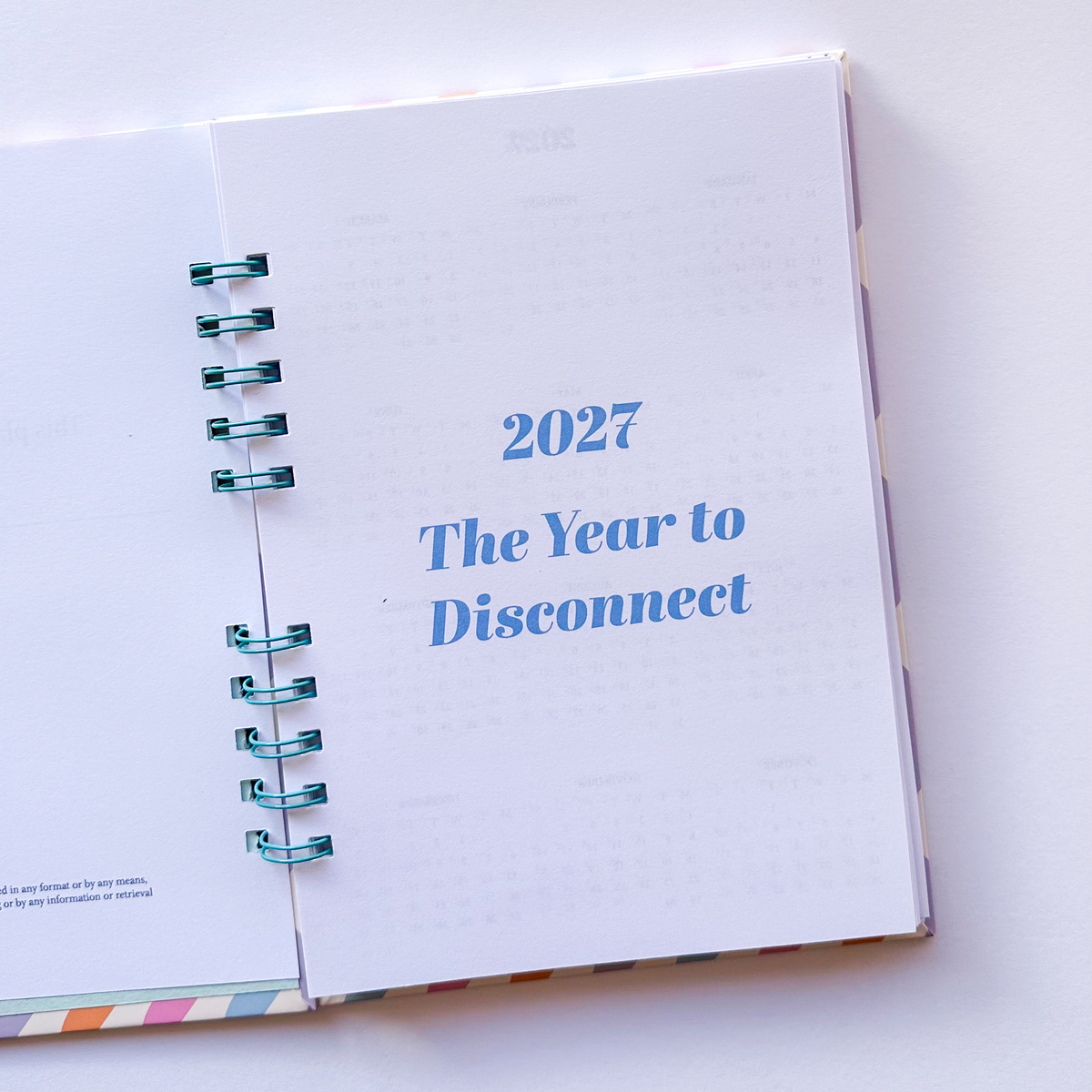 An open spiral-bound planner or notebook on a white surface, showing a page with the text '2027 The Year to Disconnect' in blue. The spiral binding is teal and silver.