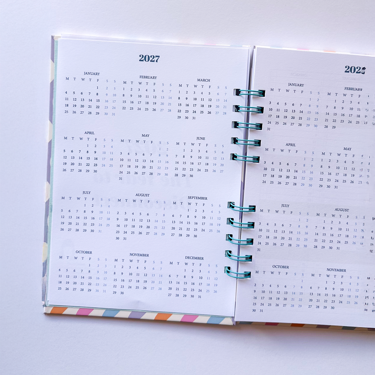 An open spiral-bound planner or calendar showing the 2027 monthly calendar pages, photographed from above on a plain white background.