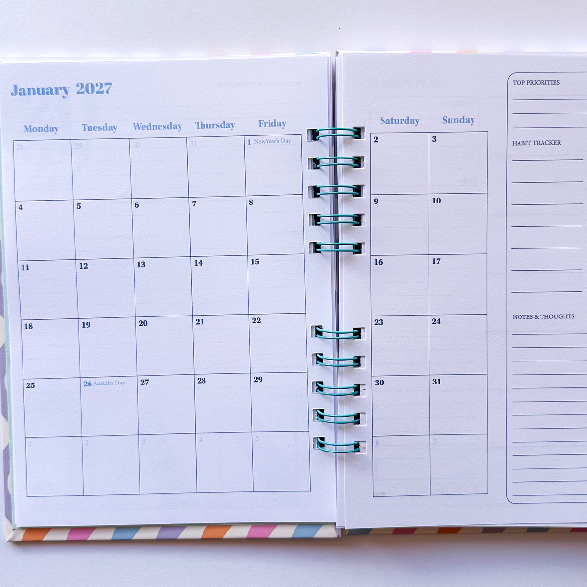 An open spiral-bound planner showing a monthly calendar for January 2027 on the left page and a weekly planner layout on the right page, with teal rings and a small multicolor striped accent at the bottom.