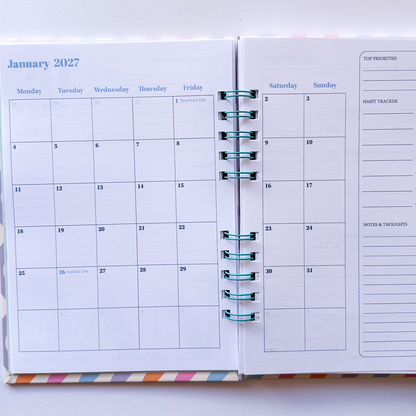 An open spiral-bound planner showing a monthly calendar for January 2027 on the left page and a weekly planner layout on the right page, with teal rings and a small multicolor striped accent at the bottom.